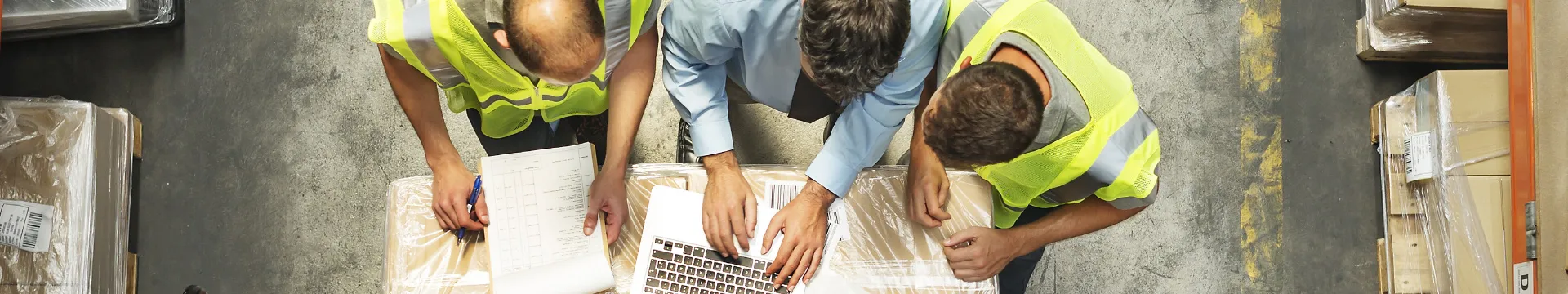 Three workers in a warehouse leaning over a box and looking at a laptop.